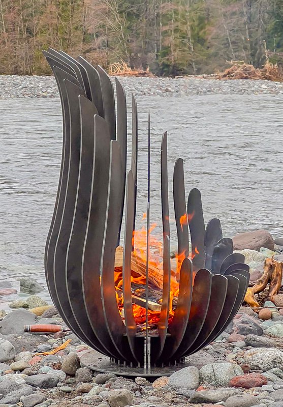 Kelray sculptural firepit burning wood on a rocky riverbank, surrounded by water and forest in a natural outdoor setting.
